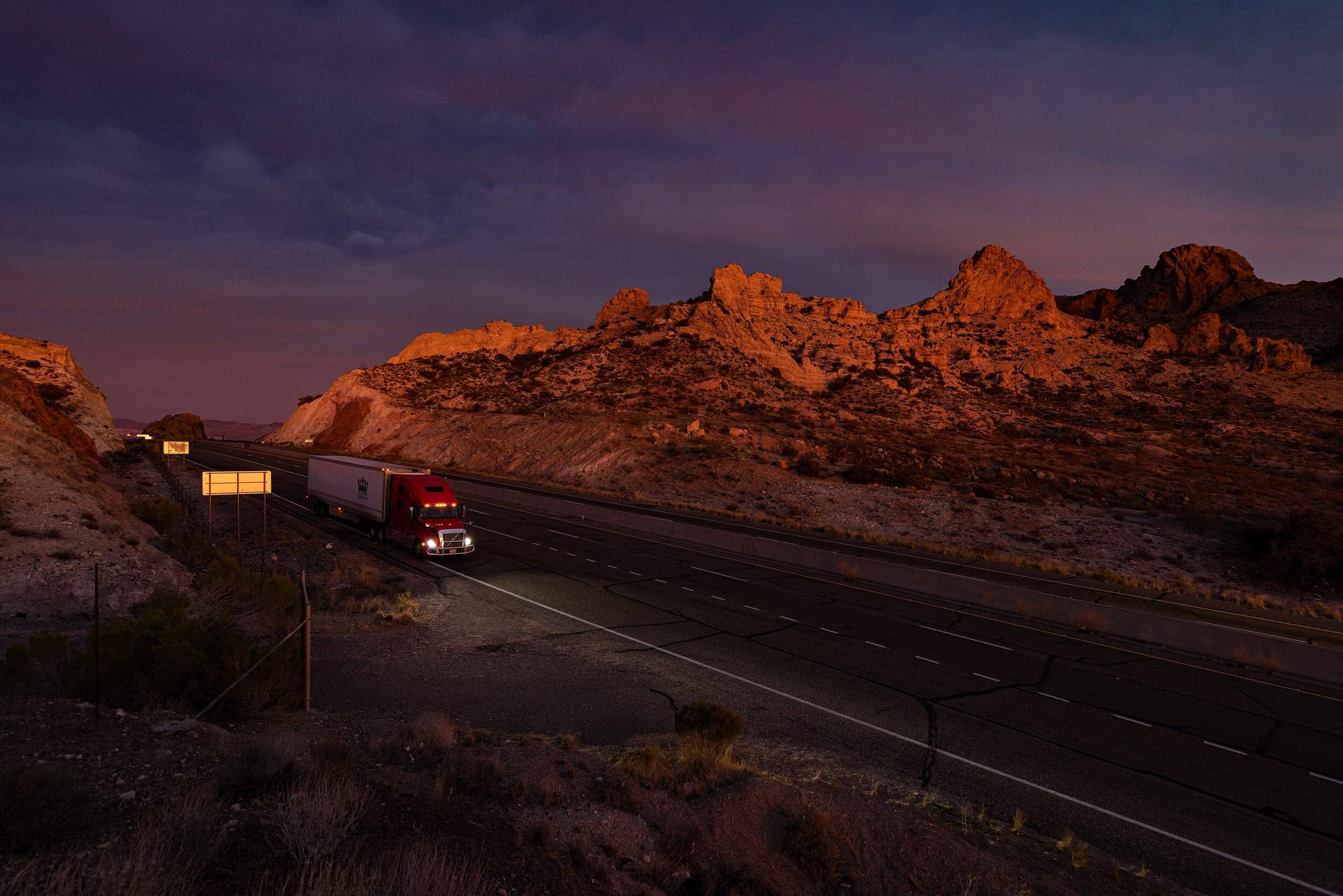 A tractor-trailer travelling a curving highway through desert hills at dusk