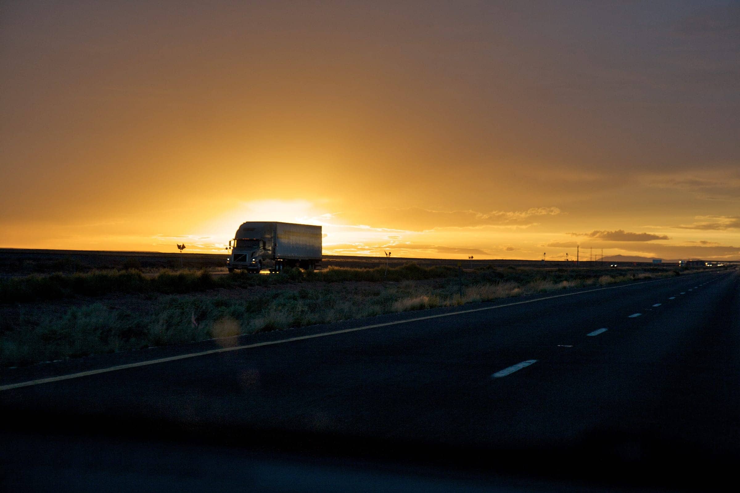 A semi-truck pulling a dry van trailer along a desert interstate at sunset