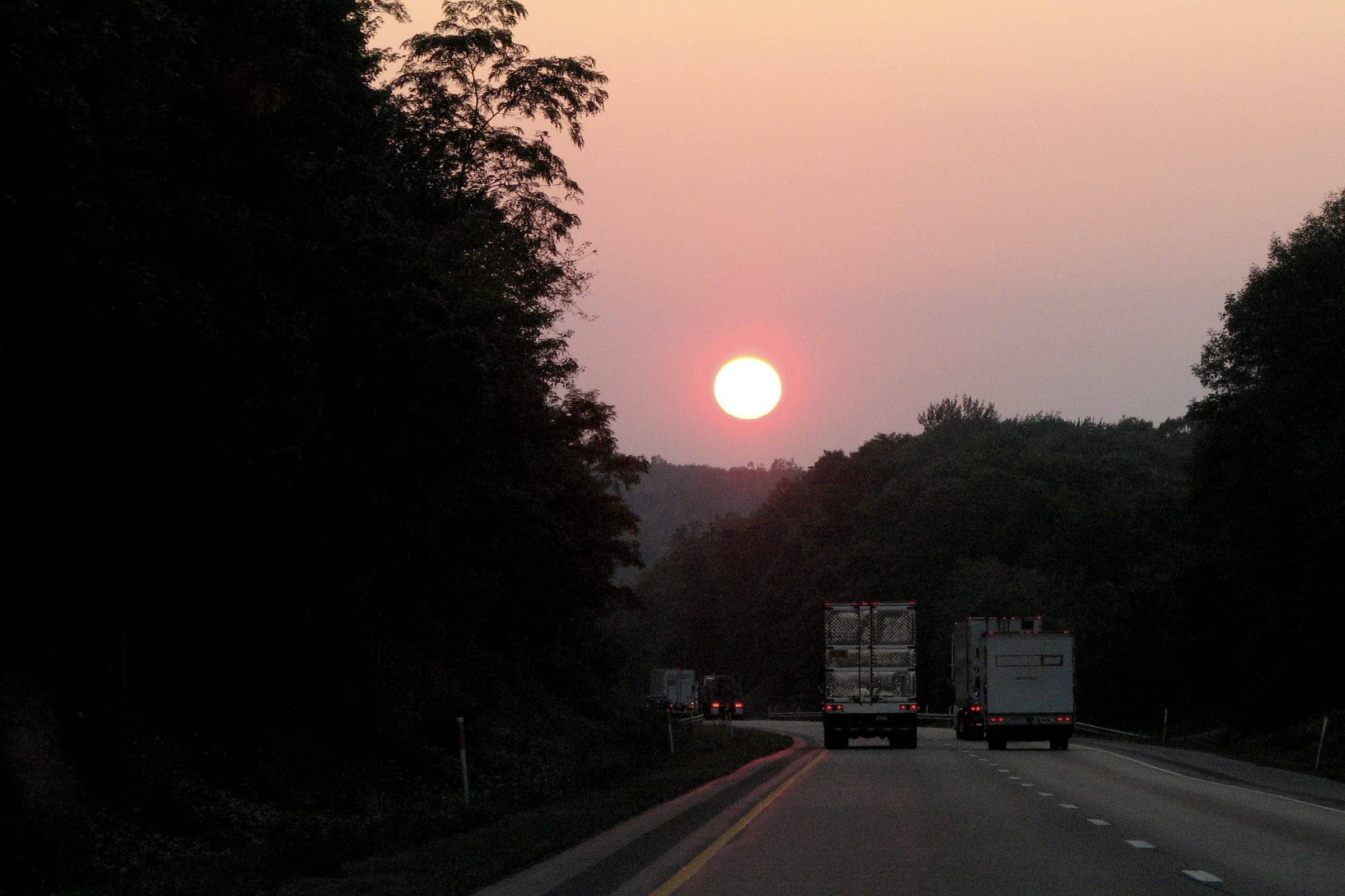 Two tractor-trailers heading down a tree-lined interstate toward the setting sun