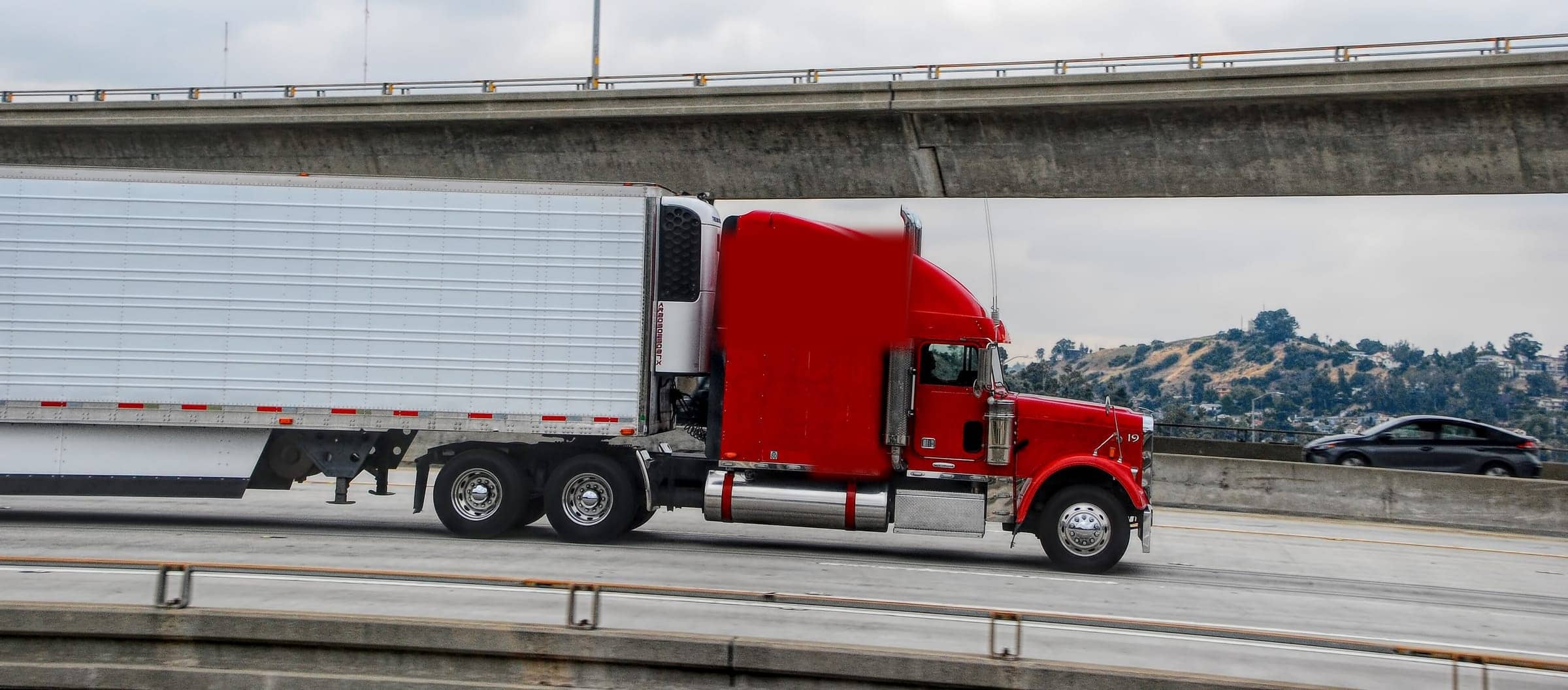 A red tractor pulling a trailer beneath a highway overpass